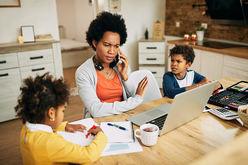 Black working mother communicating over cell phone while reading an e-mail on laptop at home.