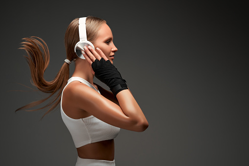 Young sport woman with headphones standing on grey background. Female boxer exercising on grey background. Side view. Copy space.