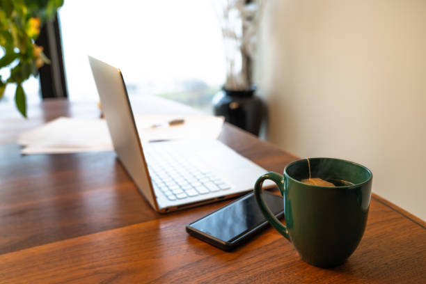 istock Work from home remote work station background image with laptop set up on a wood dining table with cell phone, papers and hot tea and light coming in from a window beyond. 1291309696