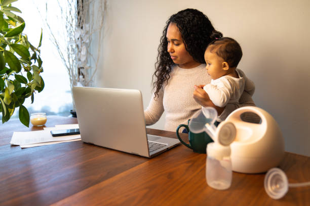 A beautiful young mixed race African American mother holds her daughter while taking notes at her dining table serving as a temporary remote work from home station with breast pump in foreground. stock photo