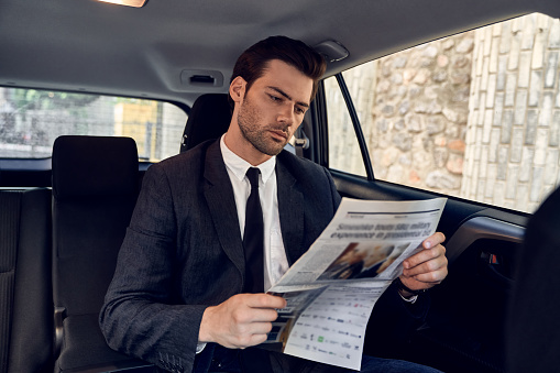 Reading latest news. Handsome young man in full suit reading a newspaper while sitting in the car.