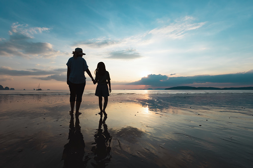 Silhouette of asian mother and daughter holding hand and walking on the beach together at the sunset time with beautiful sea and sky. Family enjoy with nature concept.