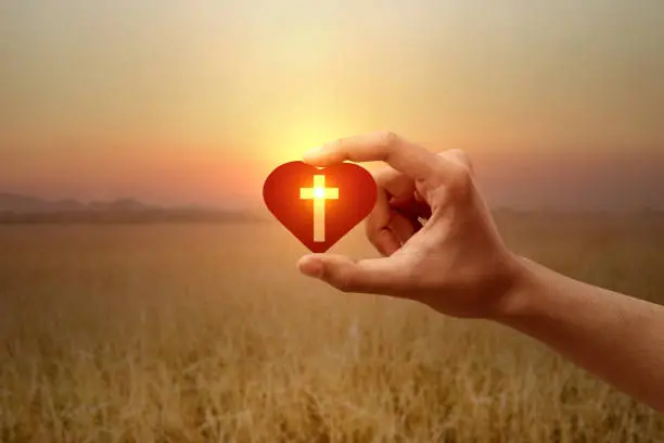Human hand holding a red heart with a Christian cross with a sunrise sky background