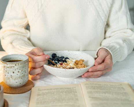 Reading book and eating healthy holiday winter breakfast with granola muesli and yogurt in bowl on white table background. Organic morning diet meal with oat