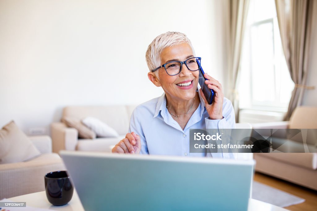 Smiling mature beautiful business woman with white hair working on laptop in bright modern home office. Business woman talking on her mobile phone while working from home Using Phone Stock Photo
