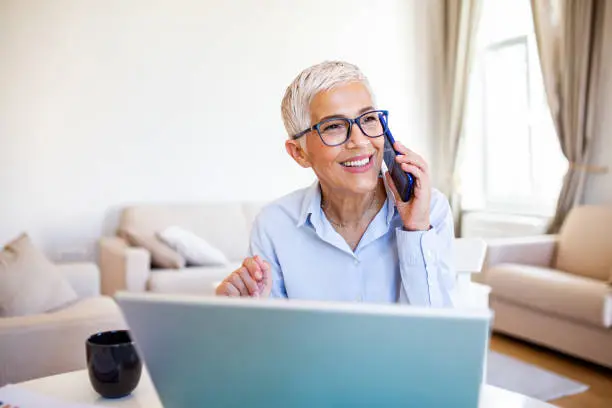 Photo of Smiling mature beautiful business woman with white hair working on laptop in bright modern home office. Business woman talking on her mobile phone while working from home