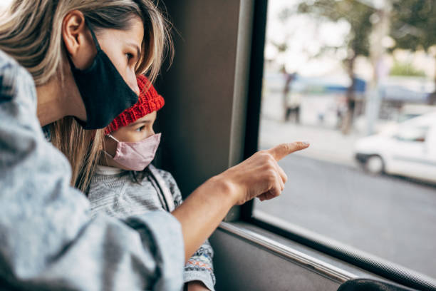 the kid wears a protective face mask in red winter hat looking into the window while going to school by bus with her mother. little girl in protective face mask looking to the route through the window - bus family travel destinations women imagens e fotografias de stock