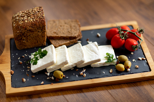 Cheese, integral bread, green olives, cherry tomato, parsley and chopped almonds on black cutting board on wooden table
