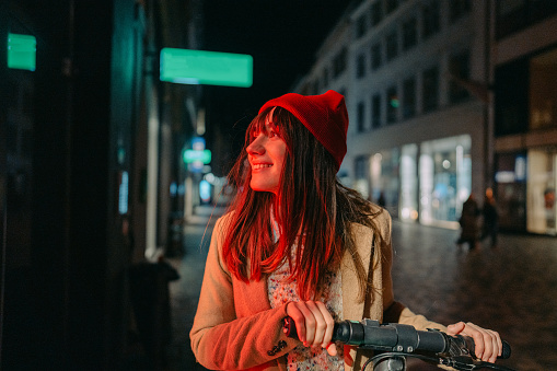 Photo of a young woman riding an e-scooter late at night, in a downtown of a city; illuminated by the neon street lights.
