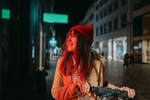 Photo of a young woman riding an e-scooter late at night, in a downtown of a city; illuminated by the neon street lights.