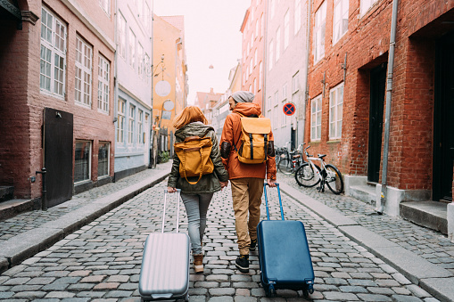 Photo of a young couple walking down the street and pulling the suitcases; exploring streets and wandering around the city; taking a trip after a long time, appreciating the view, and getting to know the new city.