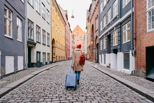 Photo of a young woman walking down the street and pulling the suitcase; exploring streets and wandering around the city; taking a trip after a long time, appreciating the view, and getting to know the new city.