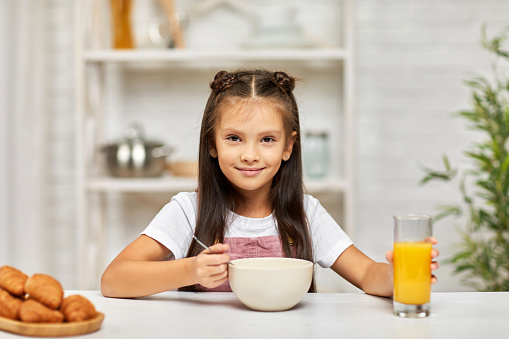 cute little child girl having breakfast - cereal and orange juice in the kitchen. healthy breakfast