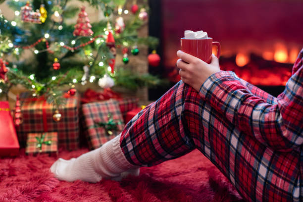 femme dans des pyjamas avec la cuve du cacao chaud et de la guimauve se reposant et se réchauffant au soir d’hiver près de la flamme de cheminée et de l’arbre de noël. - blanket fireplace winter women photos et images de collection