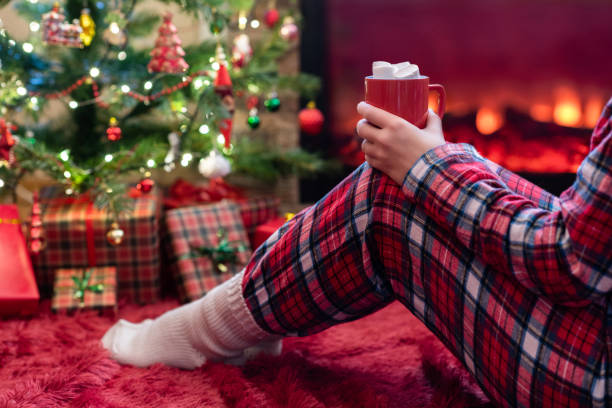 뜨거운 코코아와 마시멜로 한 잔을 들고 잠옷을 입은 여성이 벽난로 불꽃과 크리스마스 트리 근처에서 겨울 저녁에 앉아 따뜻합니다. - blanket fireplace winter women 뉴스 사진 이미지