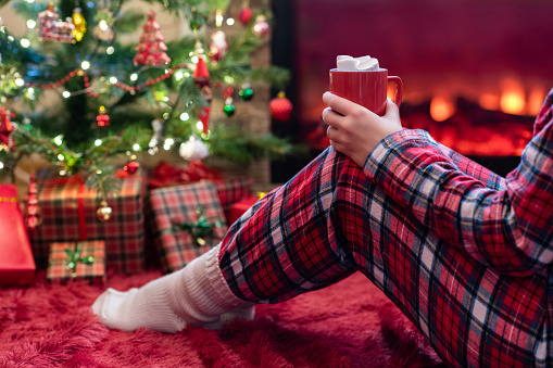 Woman in pajamas with cup of hot cocoa and marshmallow sitting and warming at winter evening near fireplace flame and  christmas tree.