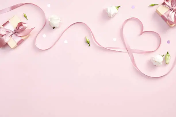 Photo of Ribbon in shape of heart with gift boxes and rose flowers on pink background. Happy Valentines day, Mothers day, birthday concept. Romantic flat lay composition.