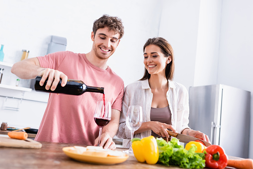 Smiling man pouring wine near girlfriend with knife and vegetables on blurred foreground