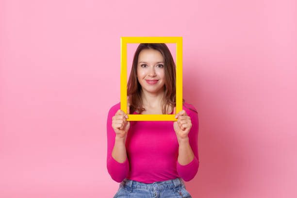 빈 노란색 프레임 안에 얼굴을 가진 젊은 유럽 여성의 클로즈업 초상화, 그녀는 그녀의 손에 들고, 분홍색 배경에 고립 - employee barracks 뉴스 사진 이미지