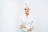 Beautiful smiling young woman has soft healthy skin after taking shower, wears bath robe and towel wrapped on head, enjoys spare time at home, isolated over white background. Wellness concept.