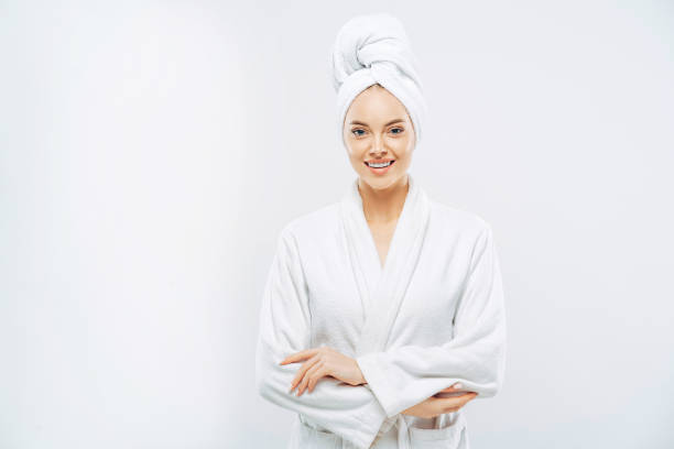 Beautiful smiling young woman has soft healthy skin after taking shower, wears bath robe and towel wrapped on head, enjoys spare time at home, isolated over white background. Wellness concept. Beautiful smiling young woman has soft healthy skin after taking shower, wears bath robe and towel wrapped on head, enjoys spare time at home, isolated over white background. Wellness concept. bathrobe stock pictures, royalty-free photos & images