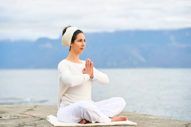 Concentrated woman meditating by the sea or lake, wearing white clothes stock photo