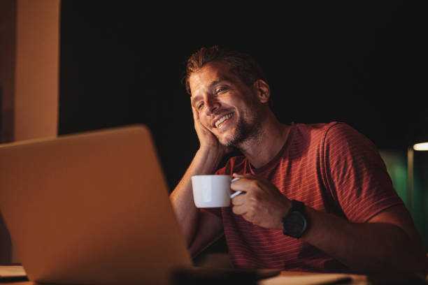 istock Handsome Happy Young Businessman Holding a Cup of Coffee while while Attending an Online Seminar at Home 1291413583