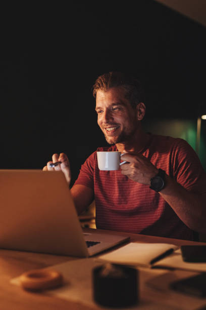 istock Handsome Happy Young Businessman Holding a Cup of Coffee while while Attending an Online Seminar at Home 1291413593