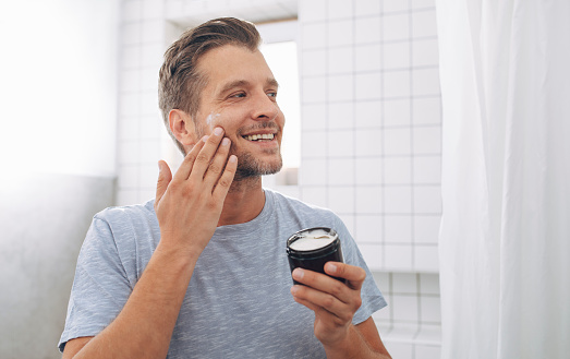 Handsome cheerful young Caucasian man applying an aftershave moisturizer after a morning shave.