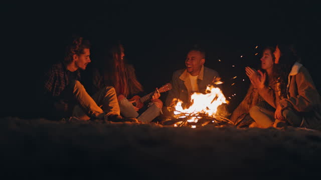 Group of friends singing playing ukulele at beach night bonfire
