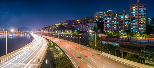 Rush hour traffic zooming along an elevated highway between the neon night cityscape and tranquil Han River in downtown Seoul, South Korea’s vibrant capital city.