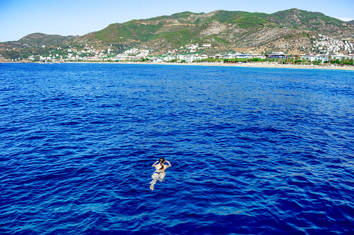 Girl swims towards the shore of Alanya (Turkey) being far from the coast in the sea. Woman lies on the surface of blue water against the background of a mountain landscape with a town on the horizon