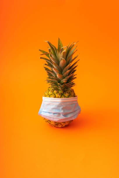 istock Pineapple with face protective mask on orange background.  COVID-19 concept. Surgical mask. Prevention 1291434552