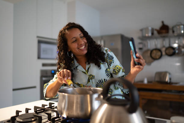 mujer adulta media haciendo una videollamada en el teléfono inteligente mientras cocina en casa - equipment housework remote domestic kitchen fotografías e imágenes de stock