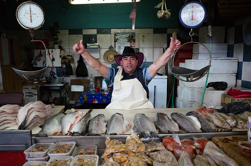 Osorno, Chile - February, 2020: Salesman sell different seafood fish mussels on local fish market. Man in hat looking at camera and show thumbs up behind market stall.