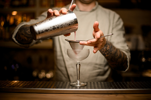 male bartender holds sieve in his hand over wine glass and pours cocktail from steel shaker cup.