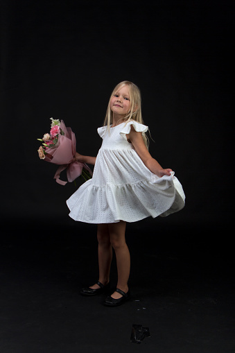 Studio portrait of cute blonde girl in white dress with beautiful gift bouquet, black backdrop, selective focus