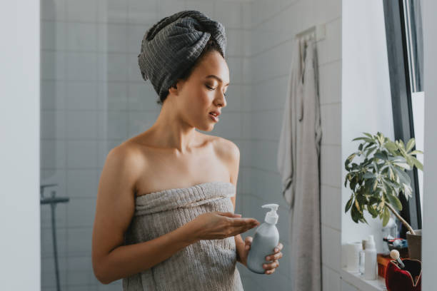 Young African American Woman Wrapped in a Towel after a Shower Holding a Skin Care Product Beautiful mixed race woman using hand moisturizer in the bathroom after a shower woman wrapped in bath towel stock pictures, royalty-free photos & images