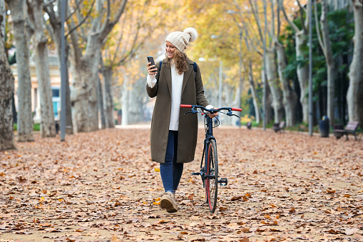 Shot of beautiful young woman sending messages with her smart phone while cycling through the park in autumn.