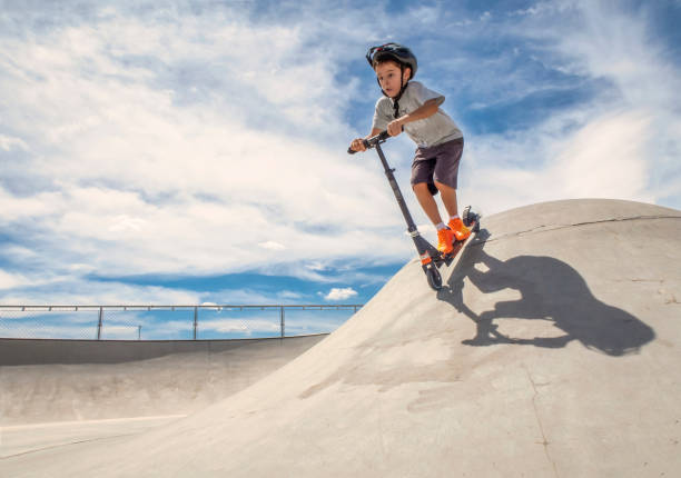 il bambino che indossa un casco scende da una rampa con uno scooter in uno skate park in una soleggiata giornata estiva, orizzontale - skateboard park foto e immagini stock