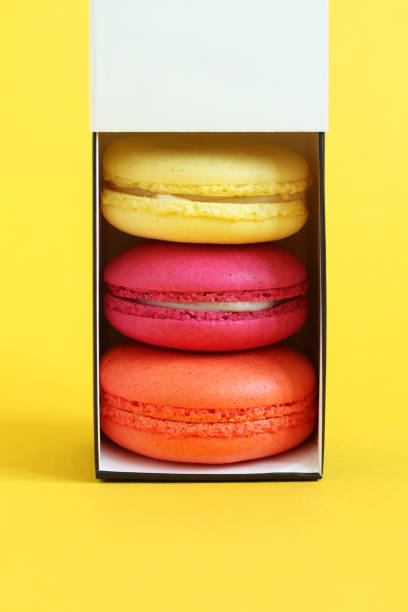 istock Multicolored macarons in a cardboard box with an open lid standing upright on a yellow background. Closeup 1291505664