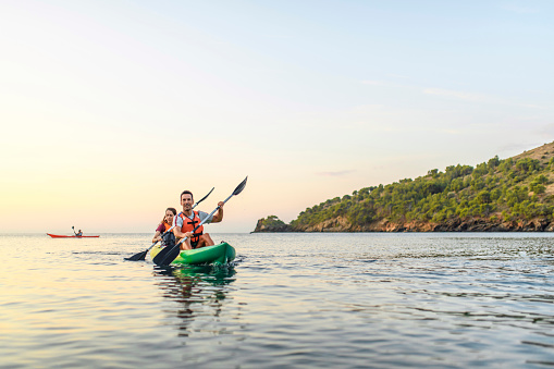 Smiling mature man and mid adult woman approaching camera as they paddle their kayak along Mediterranean coastline with single kayak in background.
