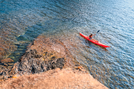 High angle view of Spanish female kayaker in early 30s enjoying healthy lifestyle as she explores rocky coastline of Mediterranean Sea.