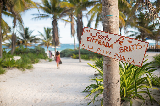 Sign on palm tree welcoming to Santa Fe beach with plants and sea as background. White and red sign with the words Santa Fe free entrance to the public beach in Spanish. Tropical summer vacations