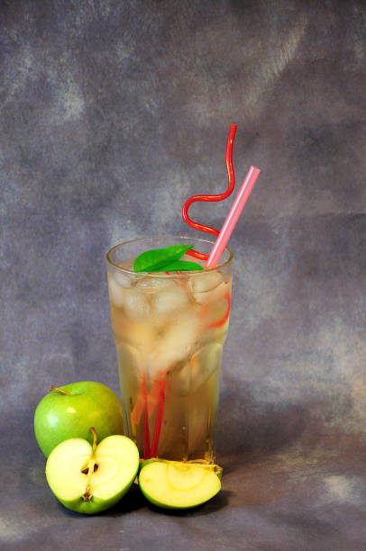 un vaso alto de una bebida de color amarillo pálido con hielo y pajitas sobre un fondo gris junto a manzanas verdes maduras. - soft cider fotografías e imágenes de stock