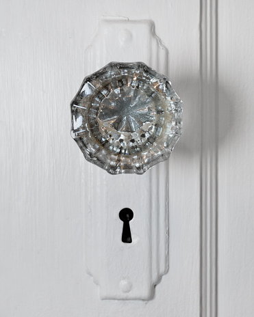 Close up photo looking straight on at an antique old crystal glass doorknob with a skeleton keyhole on a white painted metal plate and door. Copy space.
