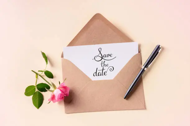 Photo of Save the date lettering on an invitation in a brown kraft envelope, shot from above on a pink pastel background