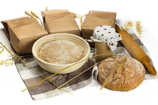 Dough in proofing basket, homemade bread, sourdough or rye leaven and paper bags of flour on the checkered table cloth. Bakery set. Craft bakery concept