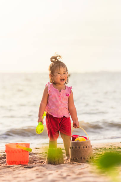 Happy fun Asian child cute little girl playing sand with toy sand tools at a tropical sea beach in holiday summer on sunset time, tourist trip concept