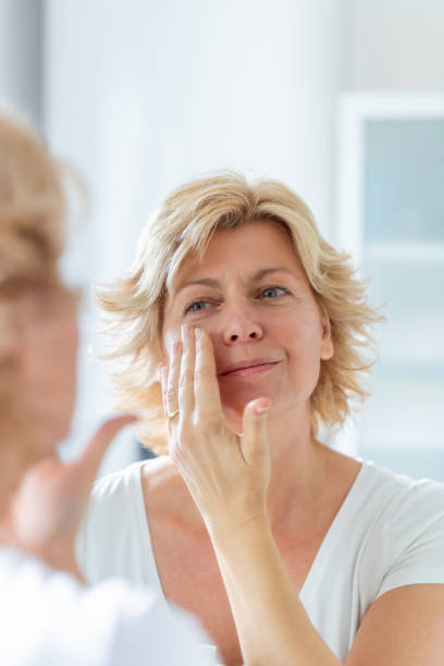 istock Blond woman's reflection in the mirror, smiling and applying face cream under eyes 1291572127